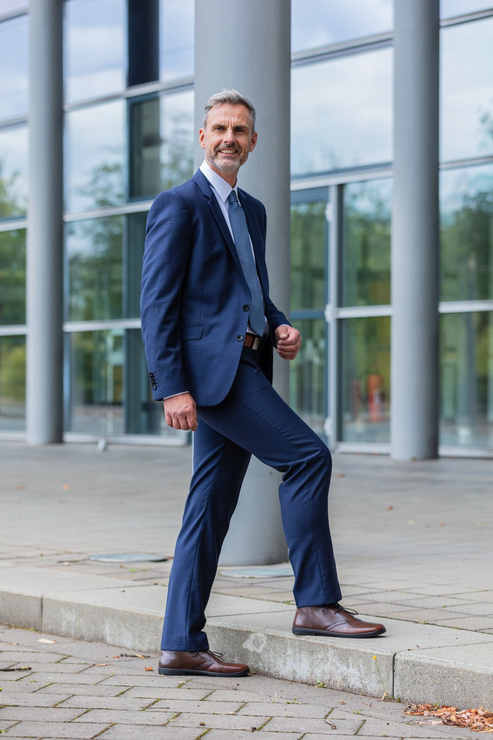 A man in a blue suit and white shirt, confidently wearing the DUQE Antique Brown shoes ZAQQ carries, stands with one foot on the same step. He stands outdoors in front of a modern glass building. The sky is partly cloudy and autumn leaves are scattered on the ground.