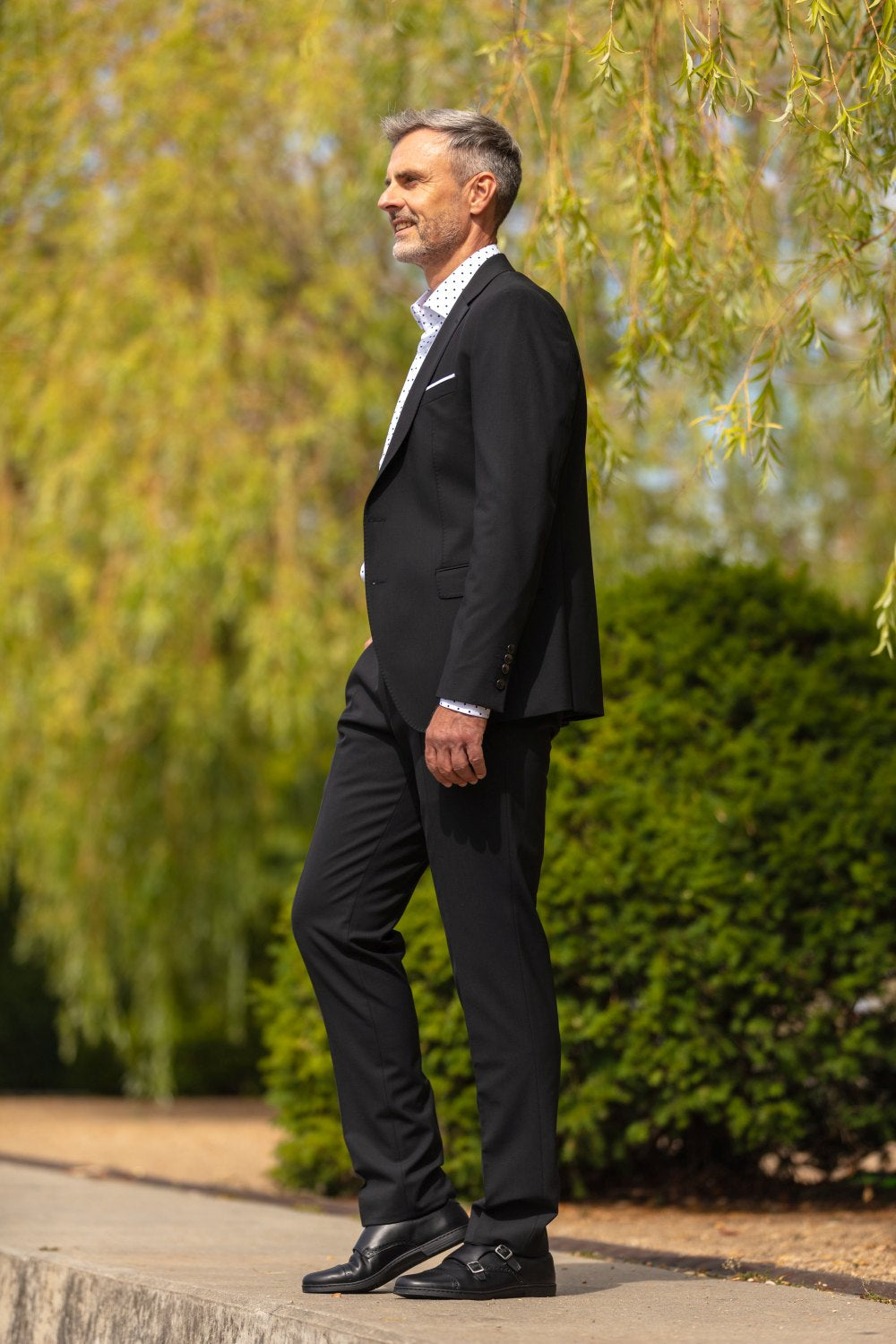 A man stands outdoors and looks to the left. He wears an elegant black suit and exudes elegance. He wears a polka dot white shirt underneath his jacket, adding a sophisticated touch to the ensemble. The scene is surrounded by lush green trees and bushes thriving under a sunny sky - a perfect backdrop to showcase the MONQ Captoe Black from ZAQQ to highlight.