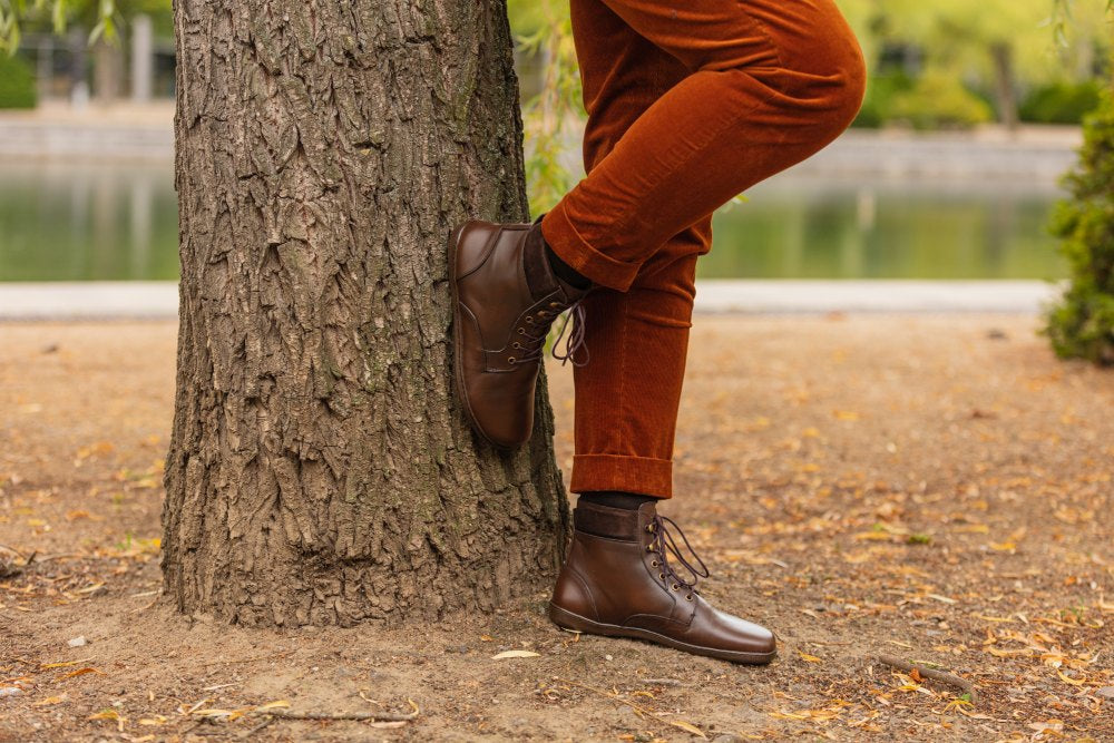 A person in QORBY brown boots from ZAQQ and rust-colored trousers leaning against a tree. A lake and green leaves can be seen in the background, reminiscent of a park landscape. The scene conveys a relaxed, autumnal atmosphere.