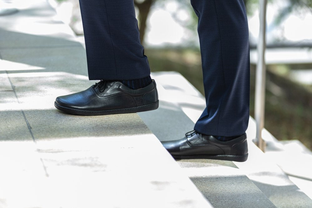 A person wearing dark blue suit trousers and black SEQUENT shoes ZAQQ climbs down a stone staircase outdoors on a sunny day. The focus is on the shoes and the lower half of the legs, while trees and sunlight form a dappled background.
