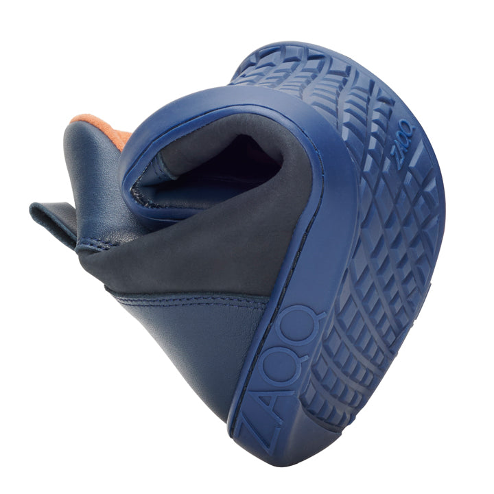 A flexible blue shoe with a thick, textured sole is shown rotated to highlight its flexibility. The soft upper underlines the streamlined design, with the brand name ZAQQ is elegantly placed next to the product name WALQER Navy Orange on the side of the sole.