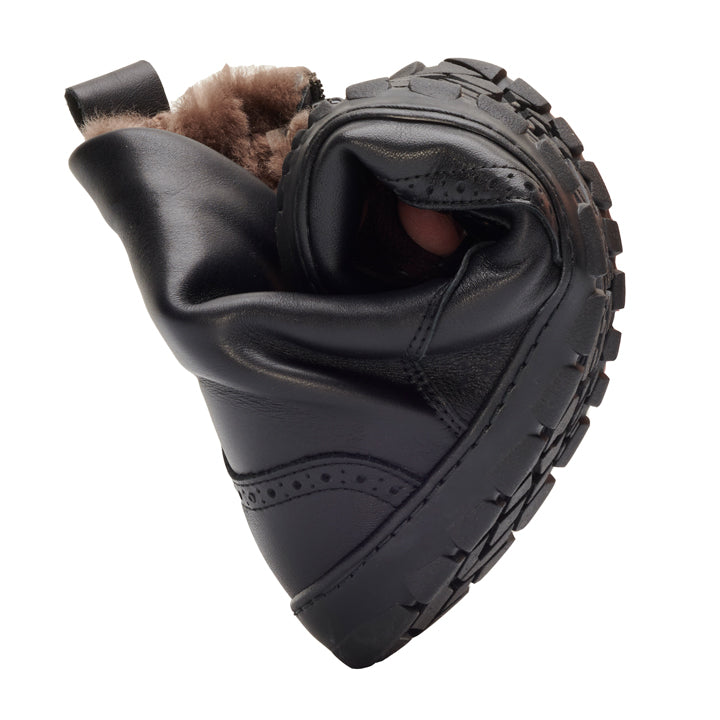 A flexible QUINTIC Winter Brogue Black from ZAQQ with sturdy sole and fur lining is twisted to demonstrate its flexibility. The shoe made from high-quality nappa leather is positioned with the fur upwards and the sole shows its structured grip pattern.