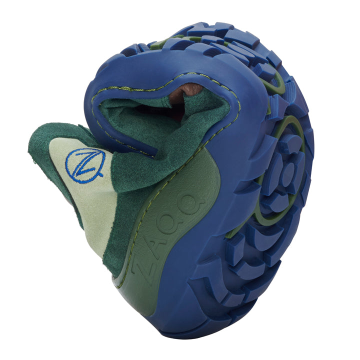 The VAQATION Green Waterproof from ZAQQ is a flexible outdoor barefoot shoe with a sturdy blue sole that is curved to demonstrate its flexibility. The upper part is green and features a logo that resembles the letter Z in blue stitching. The design emphasizes comfort, versatility and improved breathability.