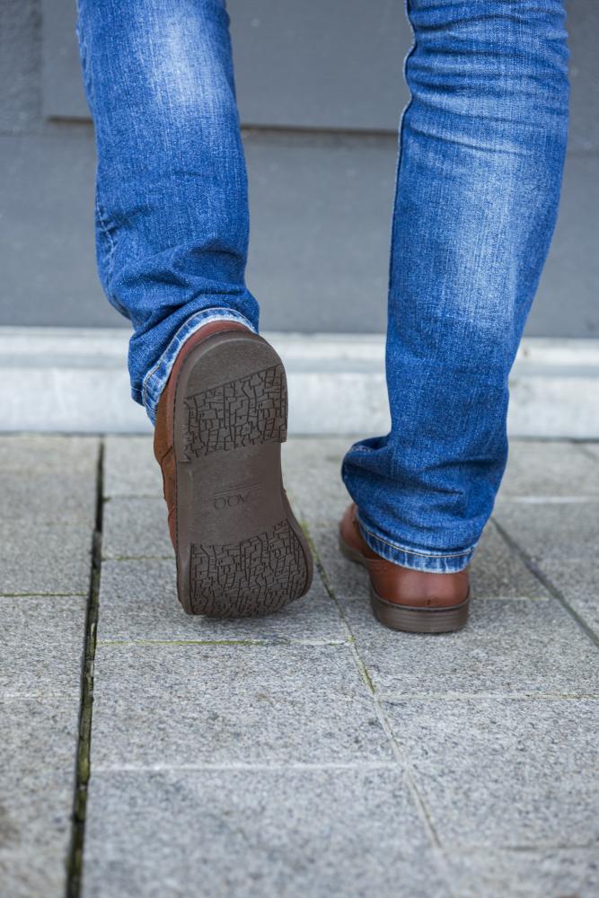 A person in blue jeans and BRIQ Brogue Antique Cognac shoes from ZAQQ runs on a tiled sidewalk, with the focus on the lower legs and the soles of her shoes.