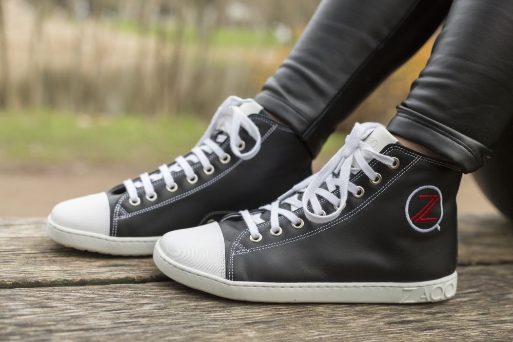 A person wearing black and white high-top sneakers with a red Z logo from the CHUQQS Black collection ZAQQ sits on a wooden bench. She is wearing black leather pants, trees and grass are gently blurred in the background.