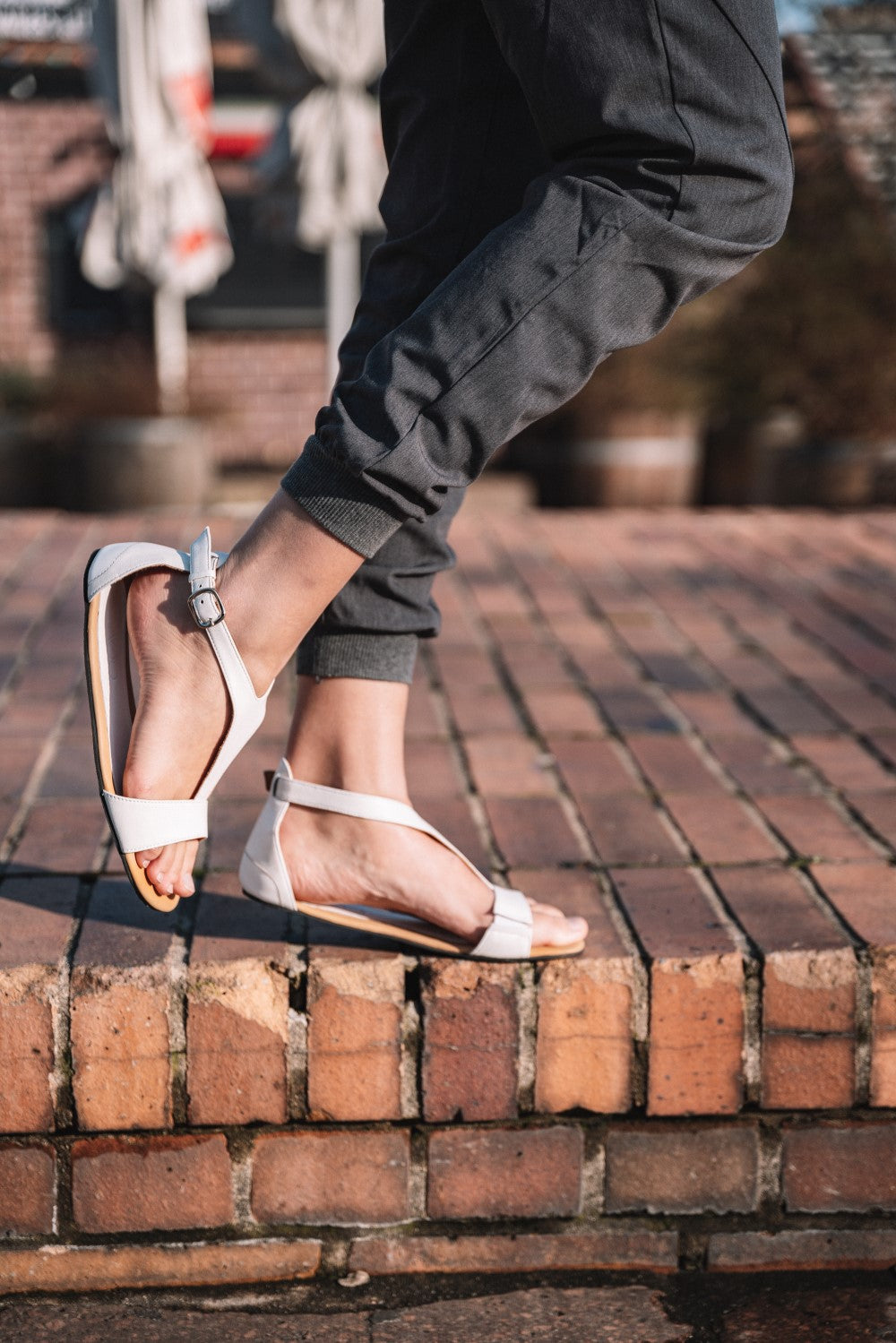 A person in rolled-up dark pants strolls on a cobblestone sidewalk wearing the flexible outsole and white barefoot sandals with ankle straps ZAQQ CLIQ.