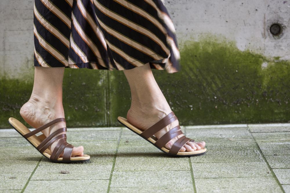 A person strolls down a green tiled path wearing the FLAQE Dark Brown Barefoot Sandals from ZAQQ and a long, striped skirt. In the background, a concrete wall with moss adds to the atmosphere, highlighting both style and exceptional comfort.