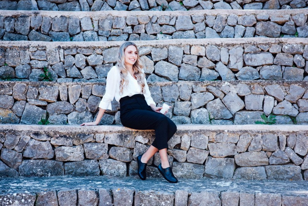 A woman with long hair, dressed in an elegant business outfit with a white shirt and black trousers, sits on a stone staircase. She smiles as she rests her right arm on the stairs and casually crosses her legs. Her polished black IQON shoes from ZAQQ add a touch of elegance to her relaxed pose.