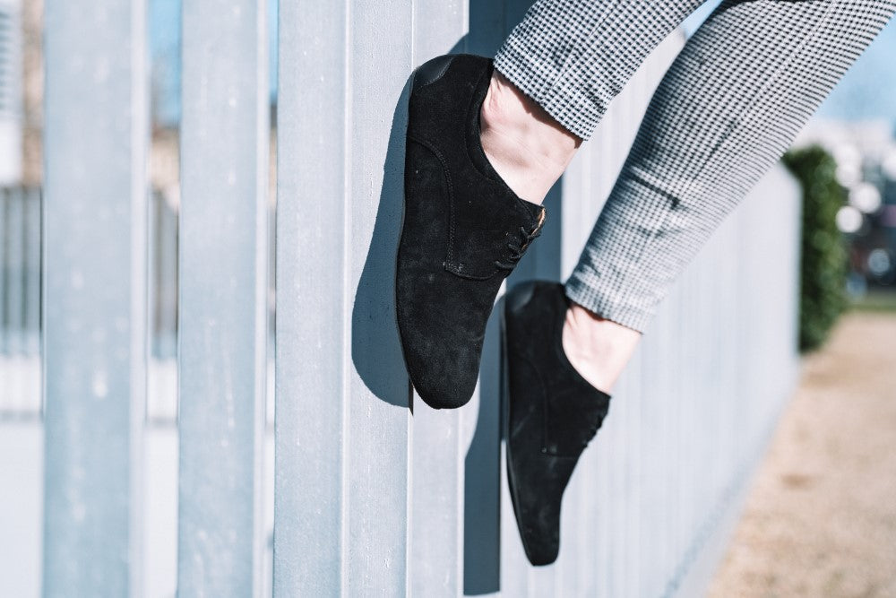 A person in black IQON velor shoes ZAQQ and checkered pants stands on a fence outdoors. The suede of the shoes harmonizes with the slightly blurred background on which grass and trees can be seen.