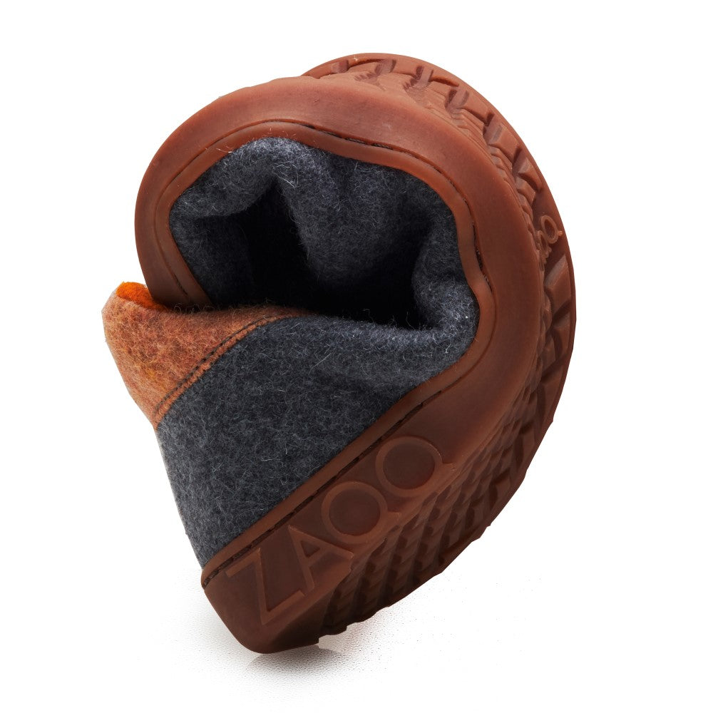 A close-up of the LIQE Gray Toffee barefoot shoe shows a brown rubber sole that is curved to demonstrate its flexibility. The upper is made of dark gray and orange textile material, with the brand name ZAQQ prominently displayed on the side.