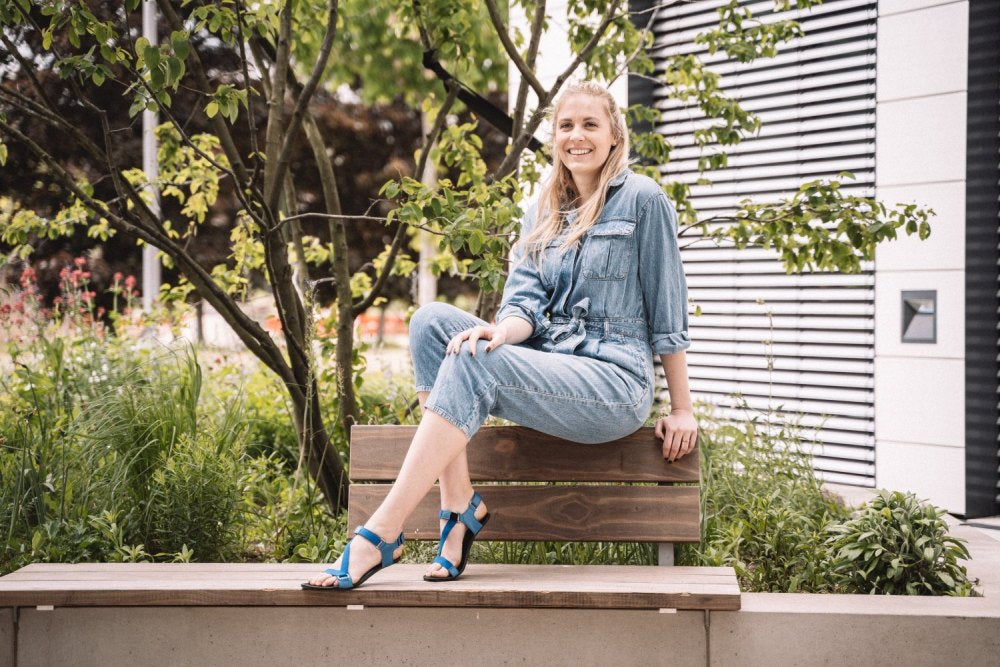 A woman sits on a garden bench, wearing a denim overall and blue MIQA barefoot sandals with durable rubber soles ZAQQ. She smiles in the greenery, surrounded by trees and bushes.