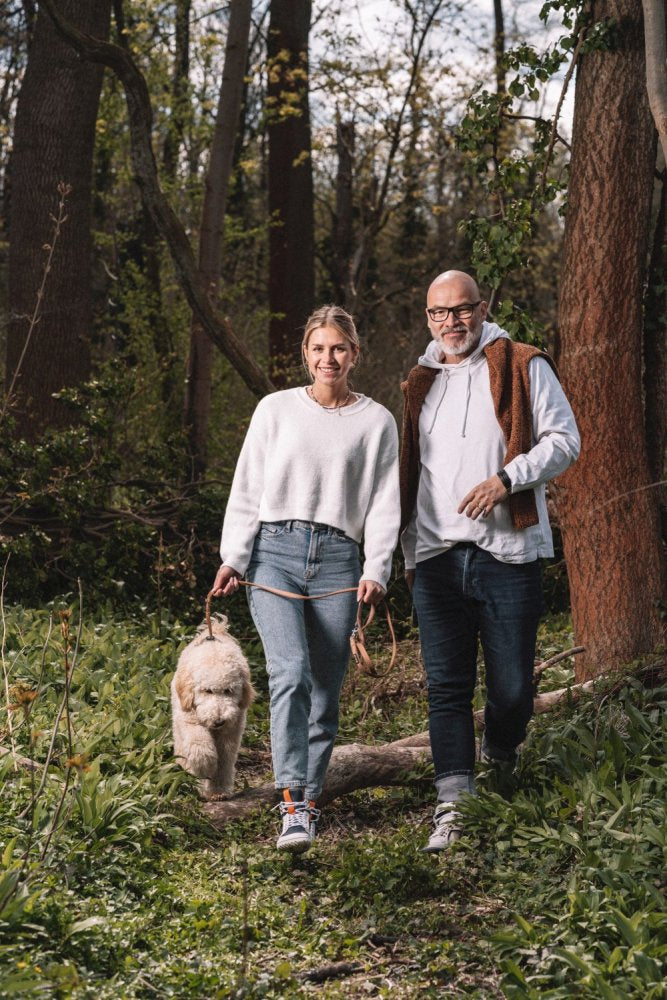 A man and a woman stroll through a forest with a fluffy dog. The woman, in a white sweater and jeans, holds the leash. The man wearing the blue and white waterproof shoes for convenience ZAQQ MONTBLANQ wears, smiling as they enjoy their sunny walk together.