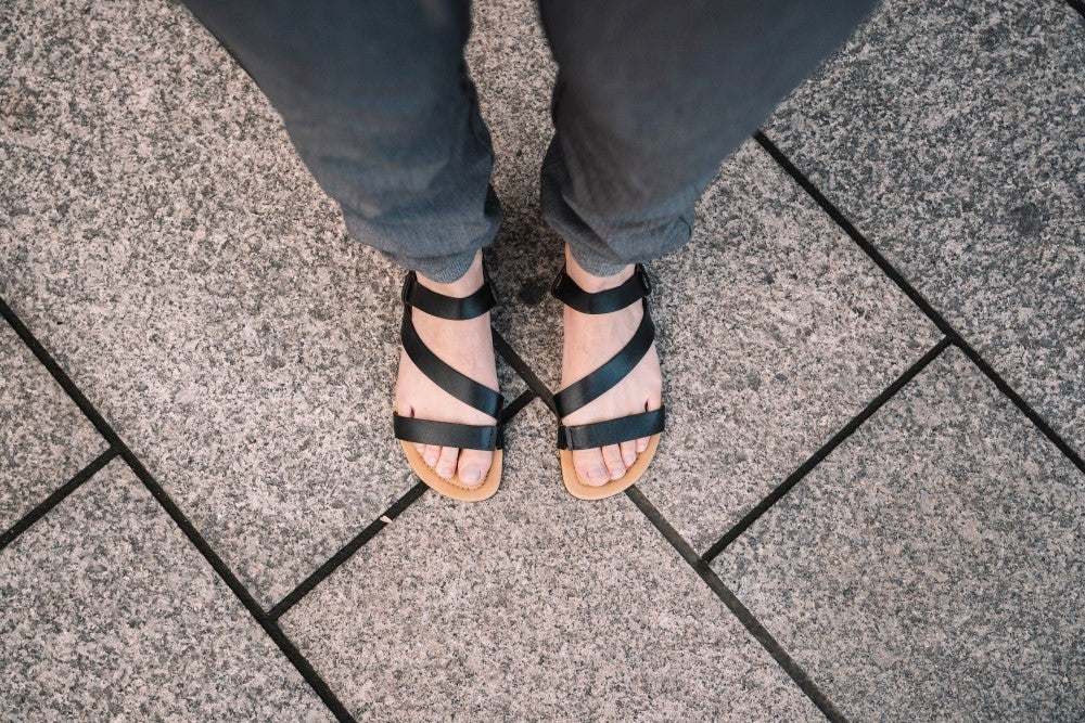 View of a person's feet in the NIQ Black sandals from ZAQQ made of high-quality nappa leather, which stands on a stone-tiled sidewalk. The elegant black strap design offers maximum comfort and perfectly complements the dark trousers.