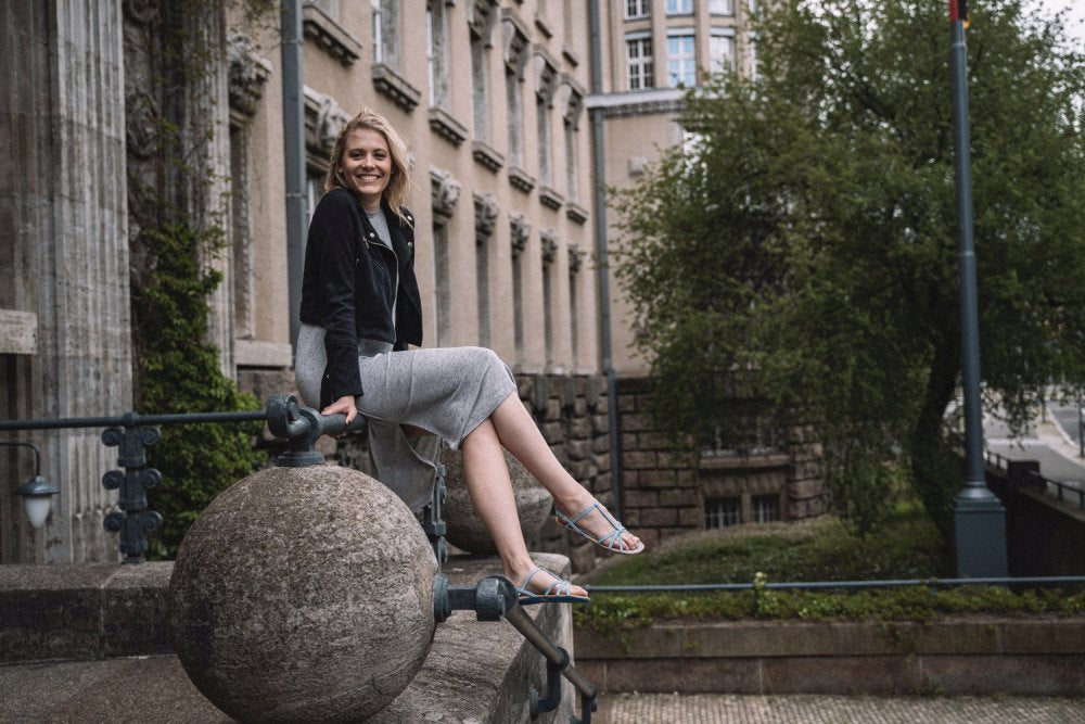 A person sits smiling on a stone railing in front of a historic building, wearing a black jacket and gray dress and QADA Blue sandals ZAQQ, which are designed for ultimate comfort. Trees and some windows can be seen in the background.