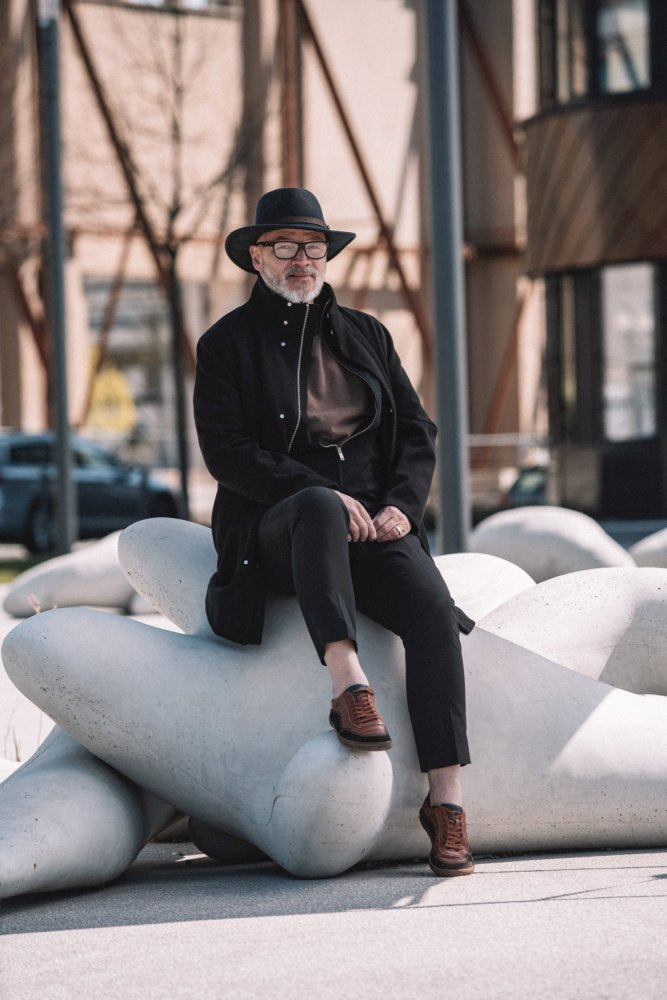 A man with a beard, a black hat, a coat and glasses sits on a large white sculpture outdoors. His dark clothing contrasts with the QAMPION Brown sneakers ZAQQ, which have flexible rubber soles. The urban background includes poles and buildings.