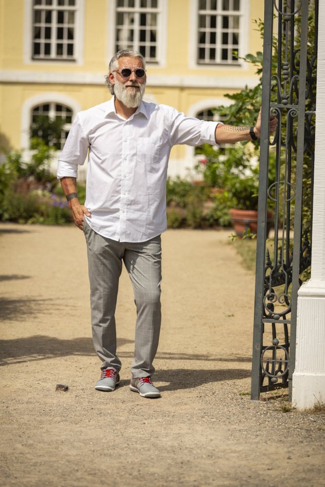 A man with a beard and sunglasses stands outside an ornate gate. He is wearing gray QANBERRA trousers from ZAQQ and a crisp white shirt. His look is completed with casual shoes made of nappa leather. In the background, a yellow building with large windows is surrounded by bright green.