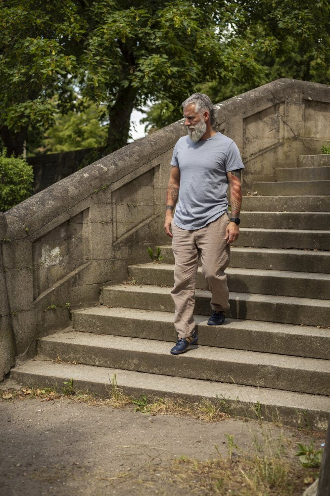 A man with a gray beard and tattoos runs down concrete steps in a park. He wears a light gray T-shirt, beige pants and ZAQQ's QANBERRA Navy barefoot shoes with minimal sole. Green trees and bushes can be seen in the background.