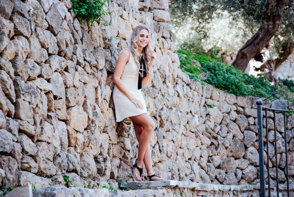 A woman in a sleeveless dress ZAQQ QAVA Black pairs it with high heels as she sits on a stone wall, smiling and adjusting her hair. She is outside amidst lush vegetation and trees, where the sunny and bright surroundings perfectly showcase her casual outfit.