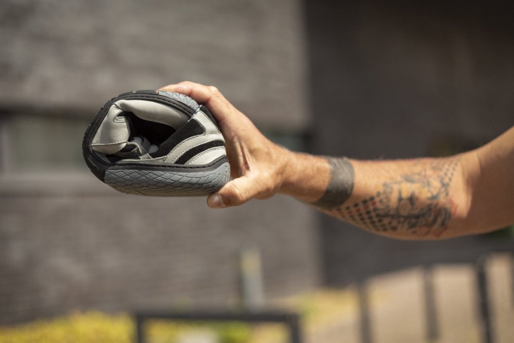 A person reaches for a pair of QENDRIQ Black Gray sneakers from ZAQQ and demonstrates her flexibility by rolling it tightly in one hand. The striped design of these barefoot shoes matches the tattoo on the wearer's forearm. A brick building comes into view in the blurred background.