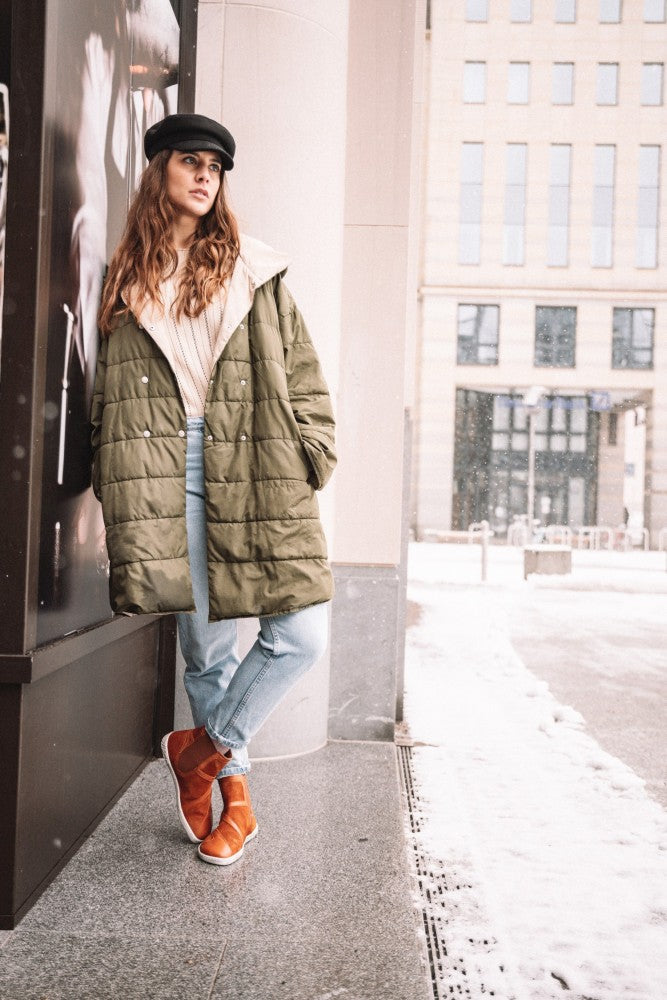 A woman stands outside in the snow, leaning against an elegant building in the color QEY WEST Cognac. She is wearing a green coat, light jeans, and orange boots ZAQQ and a black hat. The snow-covered sidewalk highlights the fresh winter landscape.