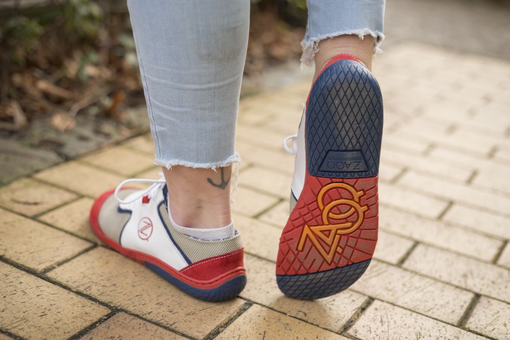 A person in rolled-up light blue jeans walks along a brick path and shows off his white QOLETTE sneakers ZAQQ. A tattoo peeks out above one ankle, while the sole of the shoe features a distinctive logo design.