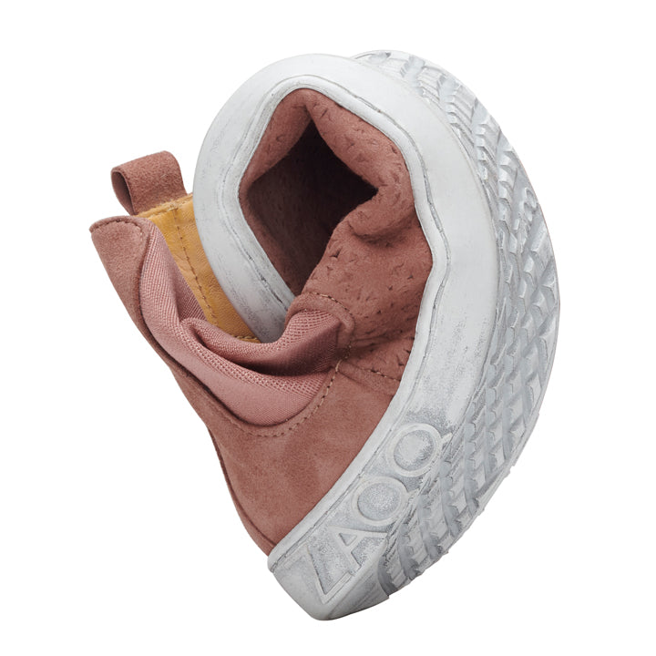 The QOLLINS Brown by ZAQQ is a pink suede baby shoe with a soft, flexible sole that rolls up to highlight its adaptability. With a light gray textured sole and a practical pull tab on the back, it is easy to wear and is ideal as a stylish barefoot shoe for little ones.