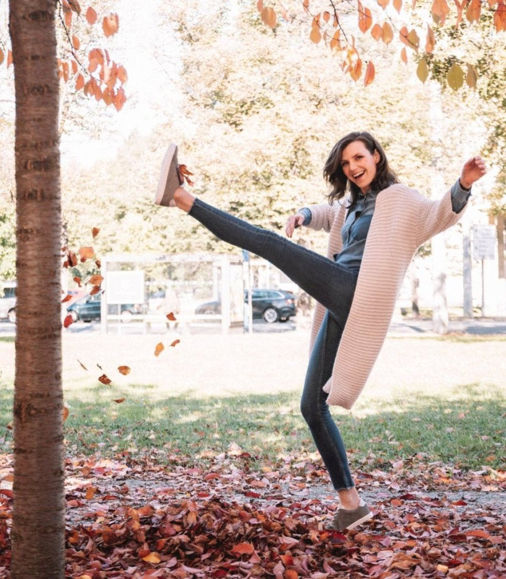 A person in a white cardigan cheerfully kicks autumn leaves while standing on a leaf-covered ground. Your QOMFY Wide Gray shoes from ZAQQ offer maximum comfort. You are in a park surrounded by trees in bright fall colors creating a bright and cheerful scene.