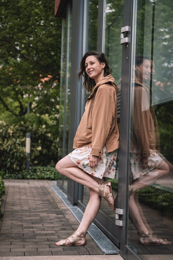 A woman in a floral dress and brown jacket leans casually against a glass wall, her smile reflected in the reflection. She stands on a brick path and shows off stylish ZAQQ QOQET Osiris sandals against the green leaf background.