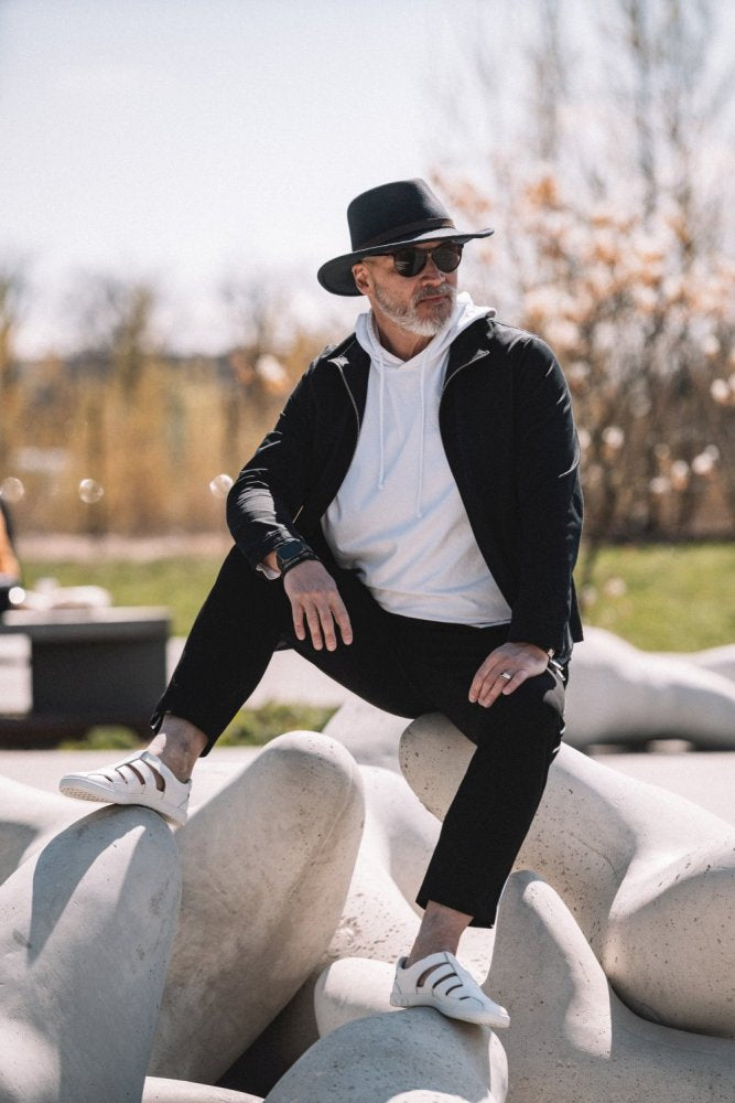 Eine Person mit schwarzem Hut, Sonnenbrille, schwarzer Jacke, weißem Hoodie, schwarzer Hose und schicken QRUISE White Sneakers von ZAQQ aus Nappaleder sitzt im Freien auf großen Steinskulpturen. Im Hintergrund sind Bäume und ein blauer Himmel zu sehen, was die ruhige Atmosphäre noch verstärkt.