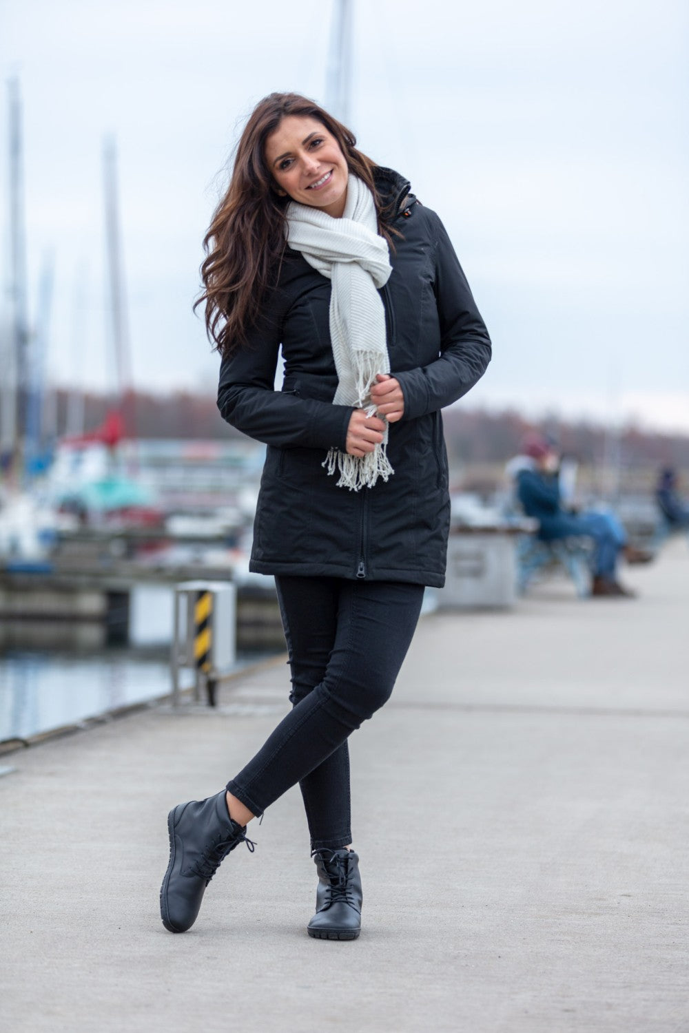 A woman stands on a sidewalk in a marina, smiling in her black jacket and pants. Your feet will fit comfortably in the QUINTIC Winter Vegan Black shoes from ZAQQ made of water-repellent material. Her white scarf provides contrast as she clasps her hands in front of her. Her long brown hair waves gently, behind her there are boats docked under the cloudy sky.