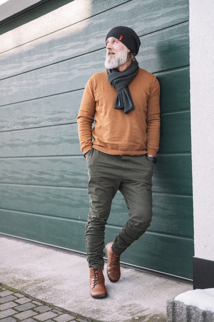 A bearded man stands outdoors and leans against a green garage door. He wears a gray knitted hat, a matching scarf, a brown sweater, green trousers and rubber-soled boots from the ZAQQ QUINTIC Winter Vegan Tabaco line. His hands are in his pockets. The floor is concrete and the scene conveys a casual winter atmosphere.