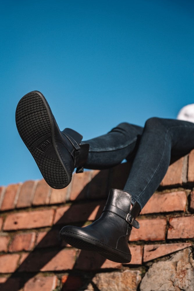 A person sits on a brick wall against a clear blue sky, wearing skinny jeans and black RIQUET boots ZAQQ. Crafted from smooth nappa leather, the dark boots dangle over the edge, giving a subtly stylish barefoot feel as their soles catch the light.