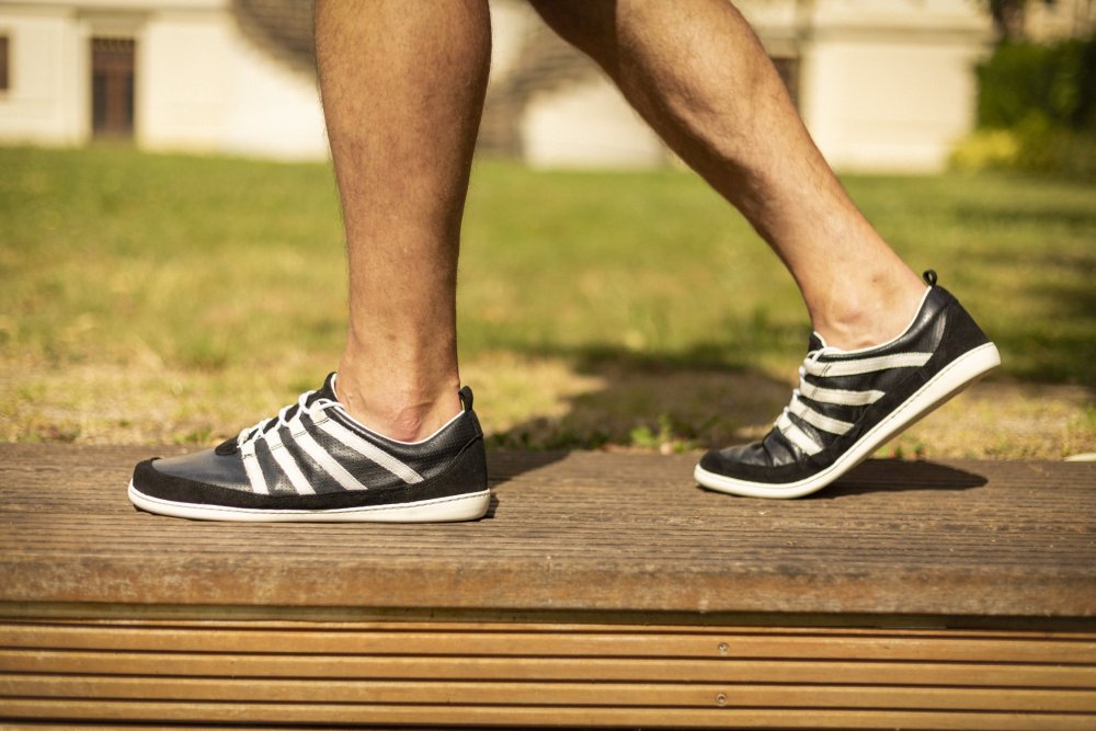 Close-up of a person walking on a wooden path wearing SPARQ Low Black White sneakers ZAQQ carries. A building is partially visible on the grassy area behind them, while only their legs and feet are visible in the image.