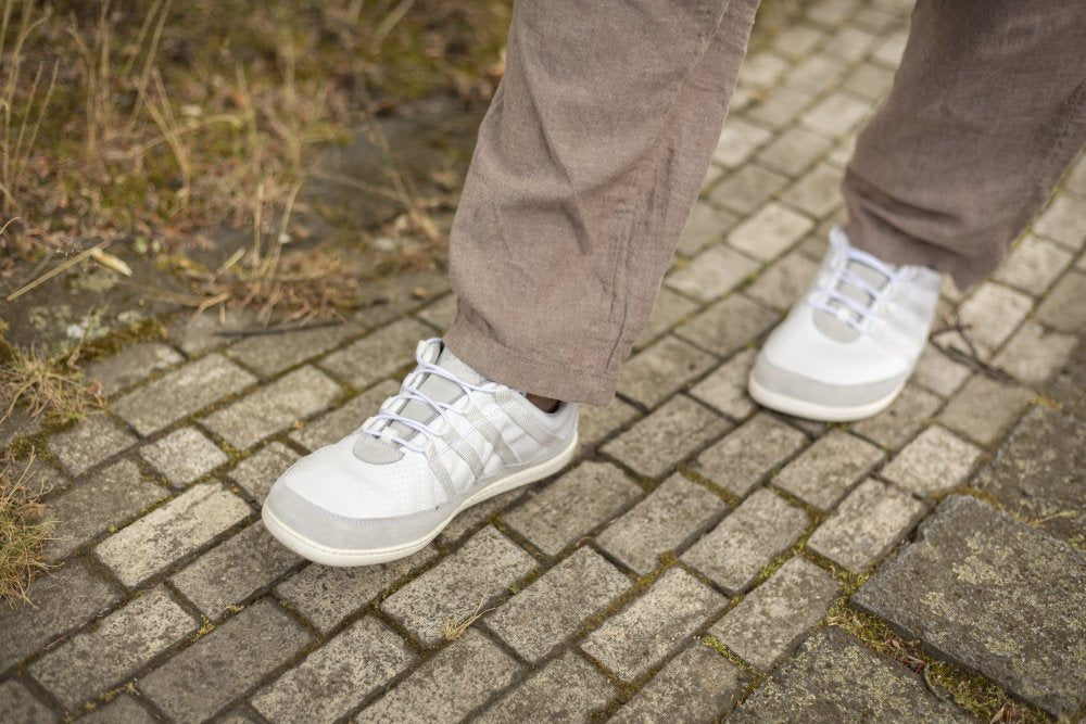 A person in gray trousers and SPARQ Low White Sneakers from ZAQQ runs on a cobblestone path. Grass and moss shimmer through the stones and enhance the outdoor atmosphere. The scene exudes a natural and casual feel, with an emphasis on comfort.
