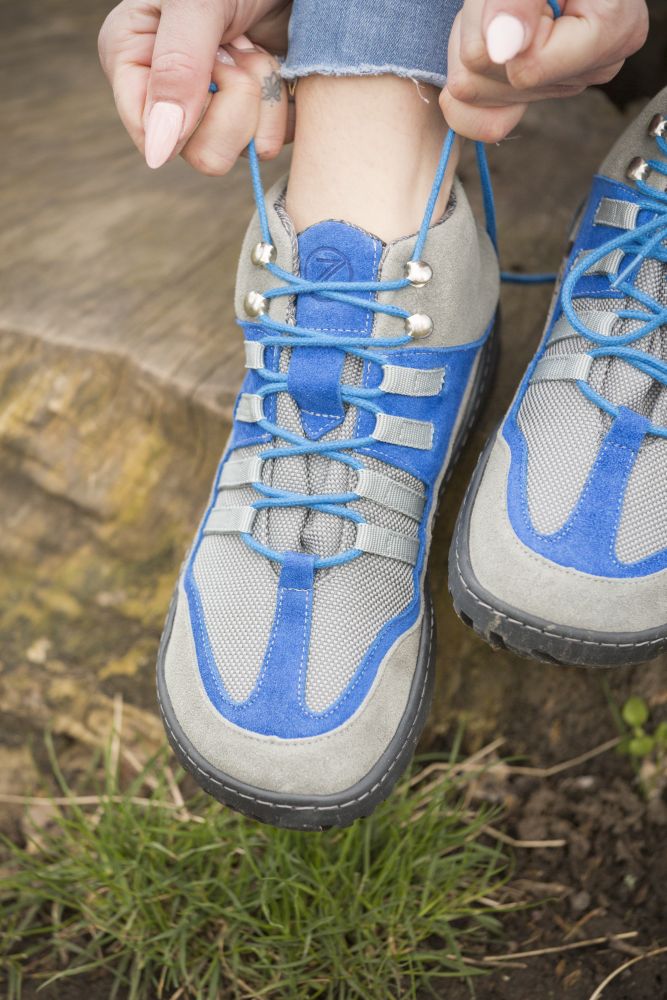 A close-up of someone sitting on a tree stump and untying the shoelaces ZAQQ SQOUT gray-blue waterproof hiking boots ties. The person is wearing jeans and grass can be seen near the stump, highlighting the breathable design that is ideal for outdoor adventures.