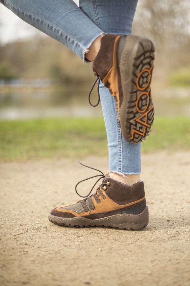 A close-up shows a person standing on a dirt path wearing the SQOUT Brown Waterproof hiking boots from ZAQQ carries. The brown and orange boots show the ZAQQ TERRA sole with visible profile. In the background, blurred greenery and water create a tranquil natural backdrop. Blue jeans complete the look.