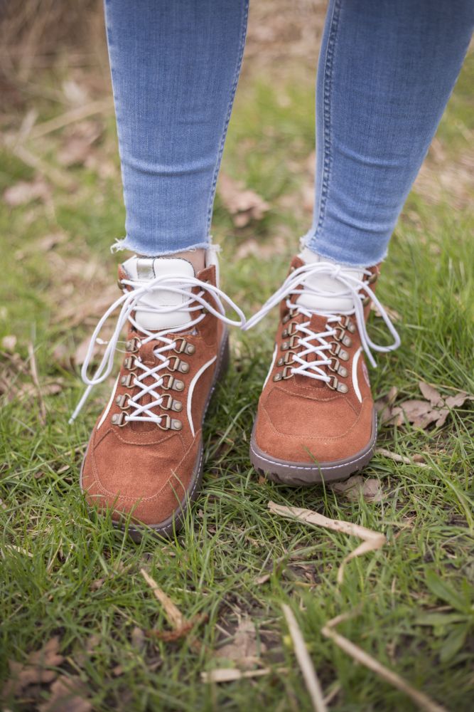 A person in blue jeans and TREQ Cognac waterproof hiking boots ZAQQ stands on green grass on which dry leaves lie scattered. The waterproof boots have white laces and metal eyelets.