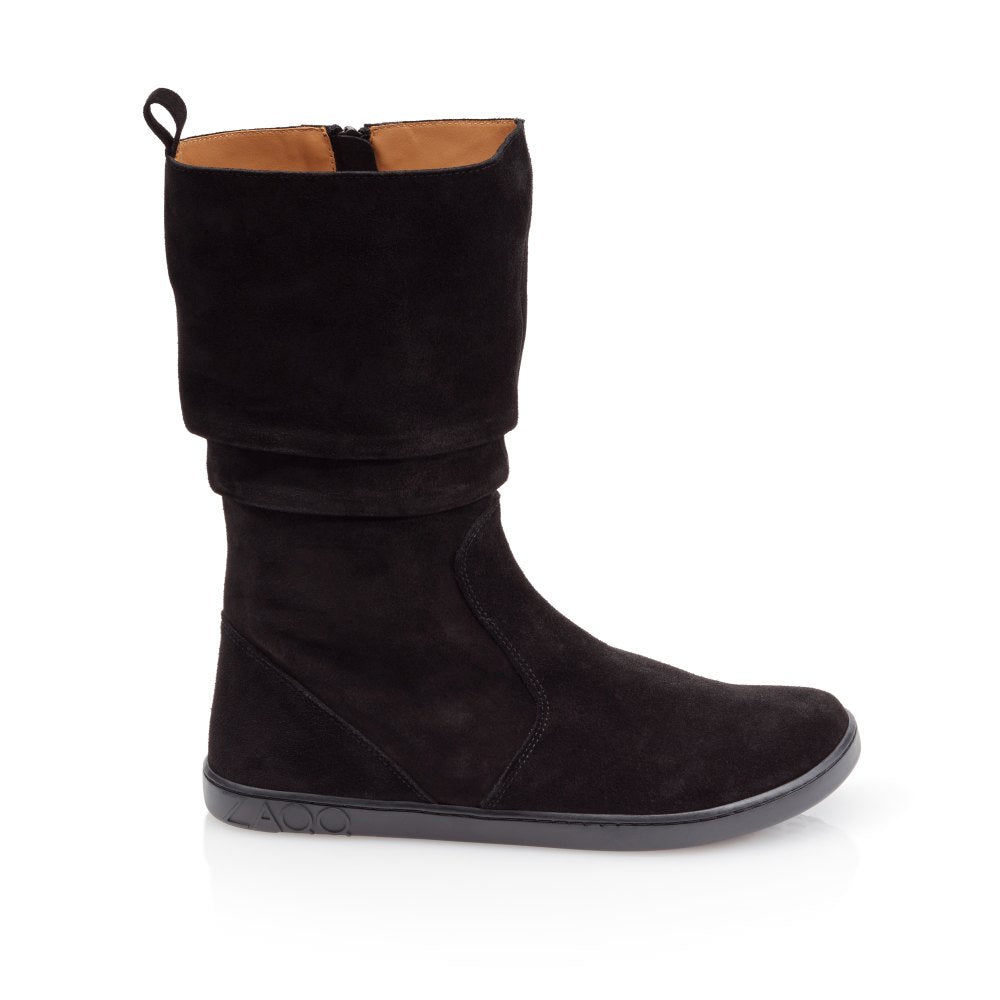 The ZAQQ QOM Black is a black suede knee-high boot with a flat sole and subtle stitching visible from the side. It has a folded cuff at the top, is made from supple nappa leather and has a zipper for closure.