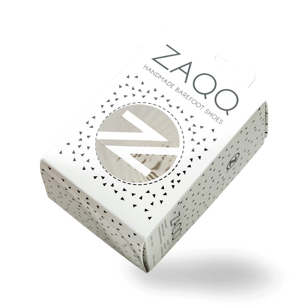 A white one ZAQQ-Box with the label Handmade Barefoot Shoes is elegantly presented. The packaging features a round transparent window that offers a glimpse of the SOQQ blue inside, accented by small black triangles scattered throughout the box.