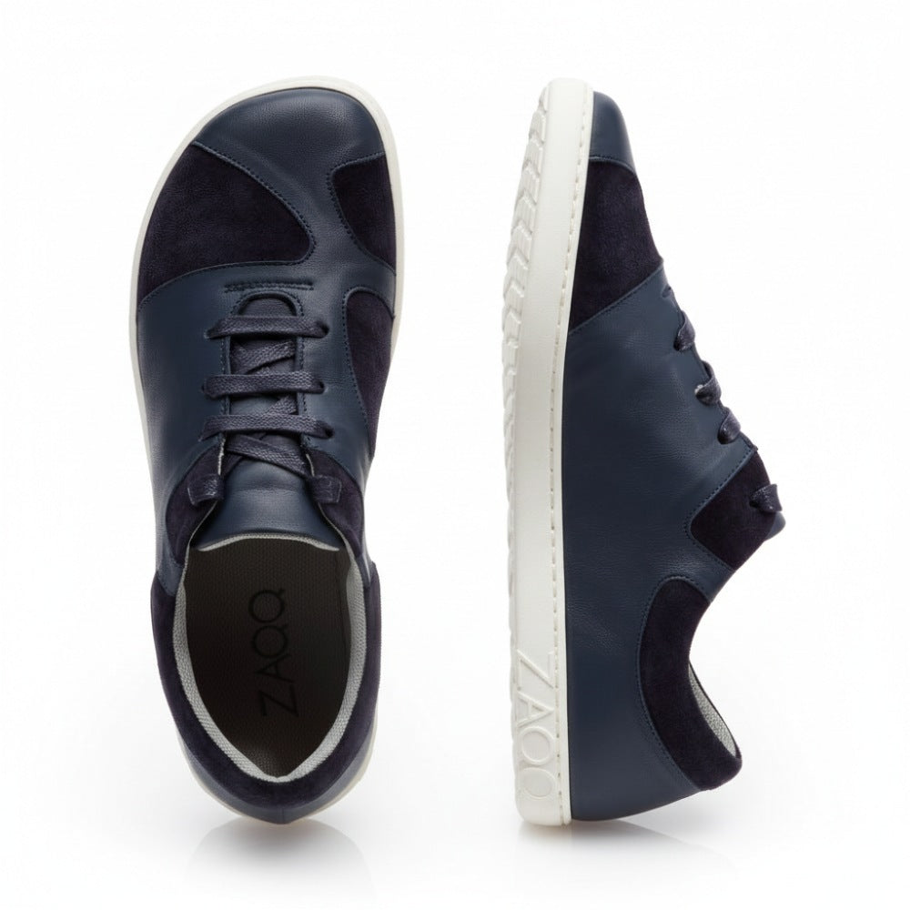 Hier werden die QAANAAQ Blue Sneaker von ZAQQ präsentiert. Ein Schuh aus elegantem Veloursleder steht aufrecht in der Seitenansicht und hat eine weiße Sohle mit dem ZAQQ-Logo. Der andere Schuh ist von oben zu sehen, was sein Schnürdesign und sein schlichtes Finish hervorhebt.