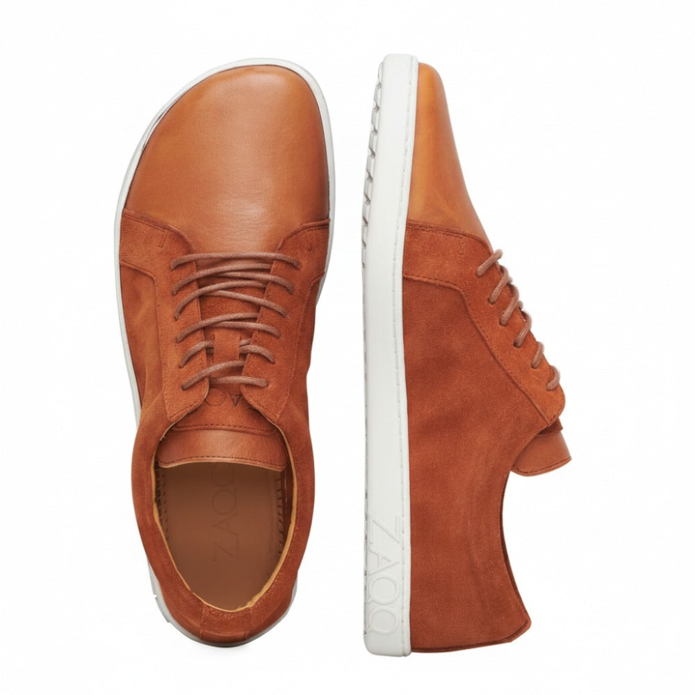 Ein Paar braune QORE Low Sierra-Sneaker wird ausgestellt, einer liegt flach, der andere steht auf der Seite. Diese stylischen Barfußschuhe von ZAQQ haben weiße Gummisohlen mit dem Markennamen auf der Seite.