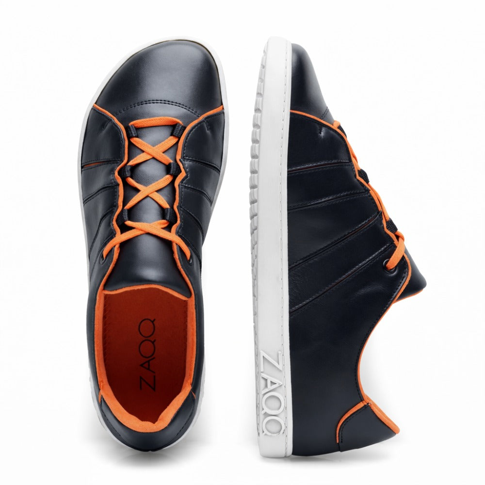 Ein Paar ZAQQ QQQ Low Black Orange Freizeitschuhe mit weißer Sohle und orangefarbenen Schnürsenkeln. Ein Schuh steht aufrecht, der andere liegt auf der Seite und trägt sowohl auf der Innensohle als auch auf der Außenseite den Markennamen ZAQQ. Diese Barfuß-Sneaker betonen mit ihrem veganen Futter einen umweltfreundlichen Aspekt.