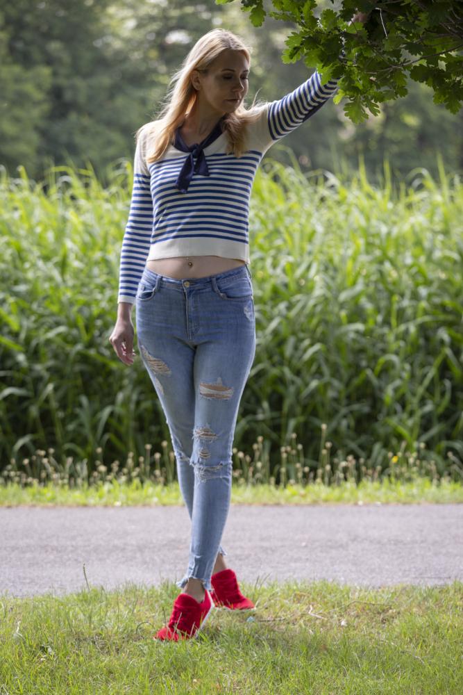 Eine Frau in gestreiftem Oberteil, zerrissenen Jeans und den LIQE Race Sneakers von ZAQQ steht auf einer Wiese und hält einen Ast. Im Hintergrund sind eine Straße und hohes Grün zu sehen. Sie sieht entspannt und nachdenklich aus.