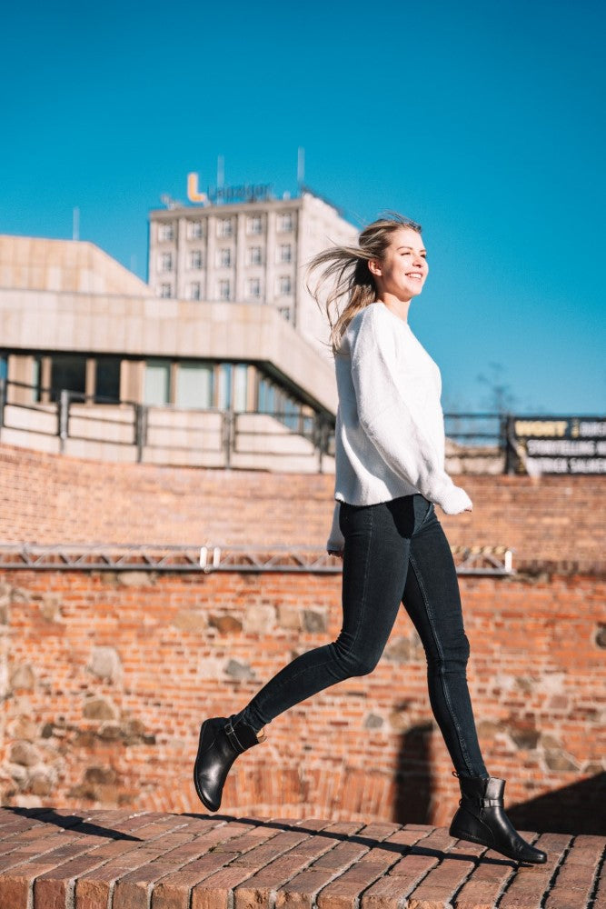 A woman in a white sweater and elegant black RIQUET boots from ZAQQ walks briskly on a brick path, her steps graceful and floating. She smiles as her hair blows in the wind and a building can be seen behind her under the clear blue sky.
