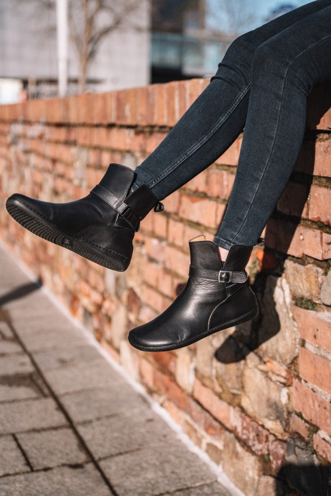 A person wears ZAQQ's black RIQUET boots made from luxurious nappa leather and pairs them with dark jeans while sitting on a brick wall. Her legs dangle happily over the edge in the sunshine, capturing the essence of the barefoot feeling with every carefree swing.