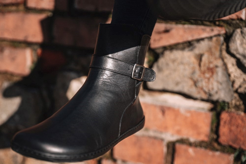 Close-up of a black RIQUET ankle boot from ZAQQ with buckled straps, worn by a person in front of a rustic brick and stone wall. The lighting highlights the smooth texture and elegant design of this stylish boot.