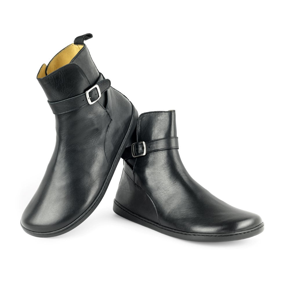 A pair of black RIQUET ankle boots from ZAQQ have elegant buckles on the sides. The left boot is slightly raised, highlighting its smooth texture and stylish design, while the right stands flat and features a polished side profile, highlighting its elegant appearance.
