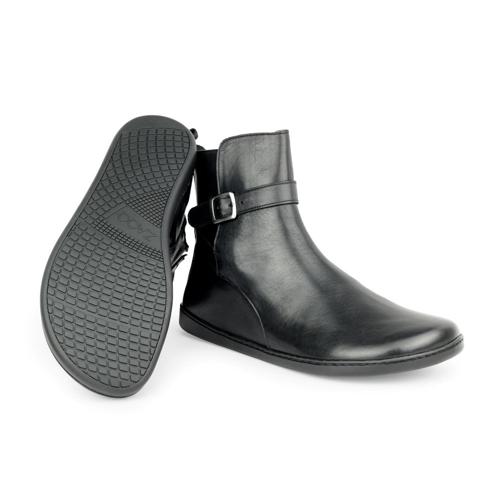 A pair of black RIQUET ankle boots from ZAQQ, one stands upright, the other shows the tread pattern of the sole. These elegant boots have a side buckle and a sleek look that offers both style and comfort.