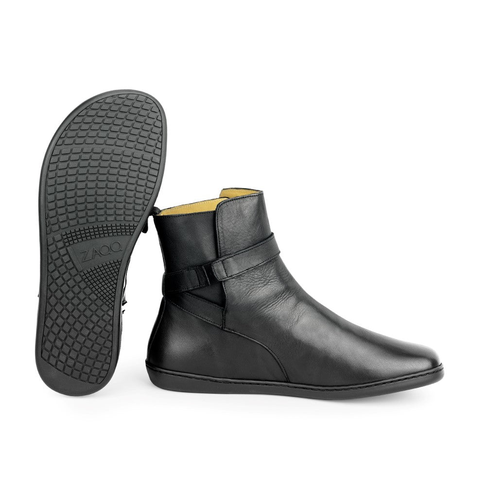 Pictured is a pair of black RIQUET ankle boots from ZAQQ. One boot stands upright, highlighting its smooth leather finish and strappy boot detail, while the other lies on its side, revealing the textured rubber sole and brand name print.